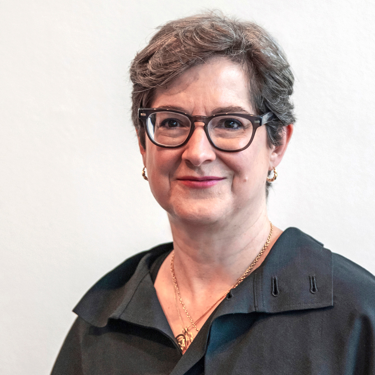 Leah Dickerman is smiling at the viewer. Dr. Dickerman has short brown hair and is wearing dark framed eyeglasses, a black blouse, and gold necklaces.