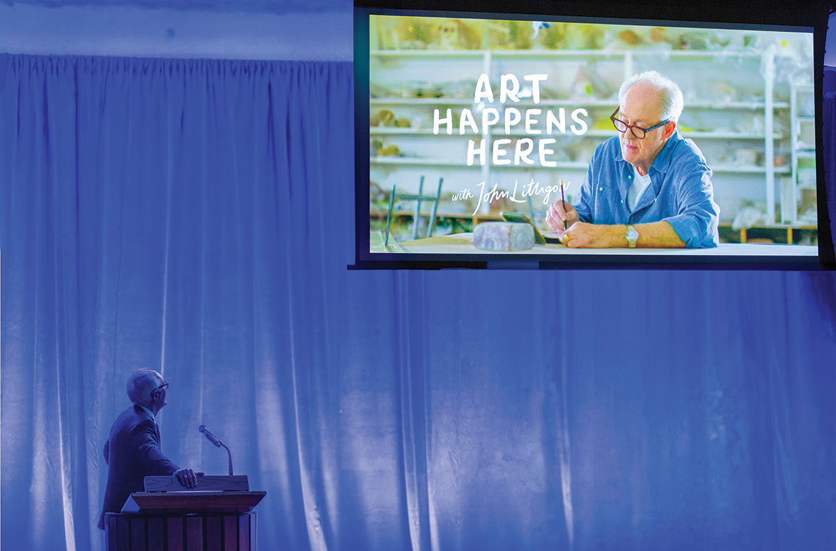 Jon Lithgow stands onstage at the 2023 Induction ceremony, looking up toward a screen where his own image is projected for the Art Happens Here project.