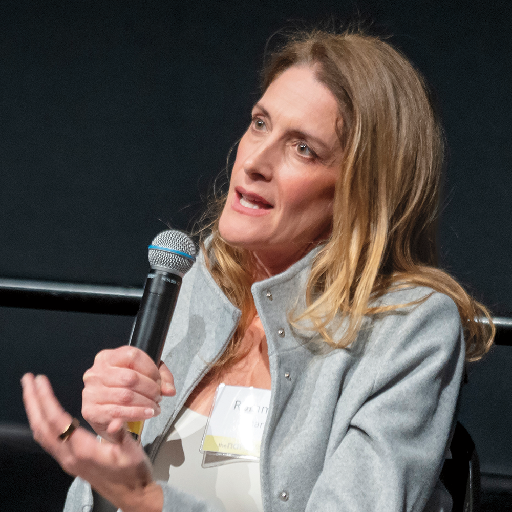 A woman with medium-length brown hair, wearing a gray jacket, is speaking into a microphone that she is holding in her hand.