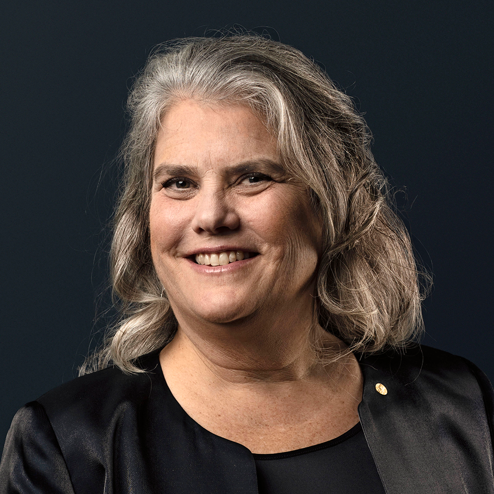 A photograph of a light-skinned individual with long gray and brown hair. They are wearing a black blazer and blouse and smiling at the viewer.  