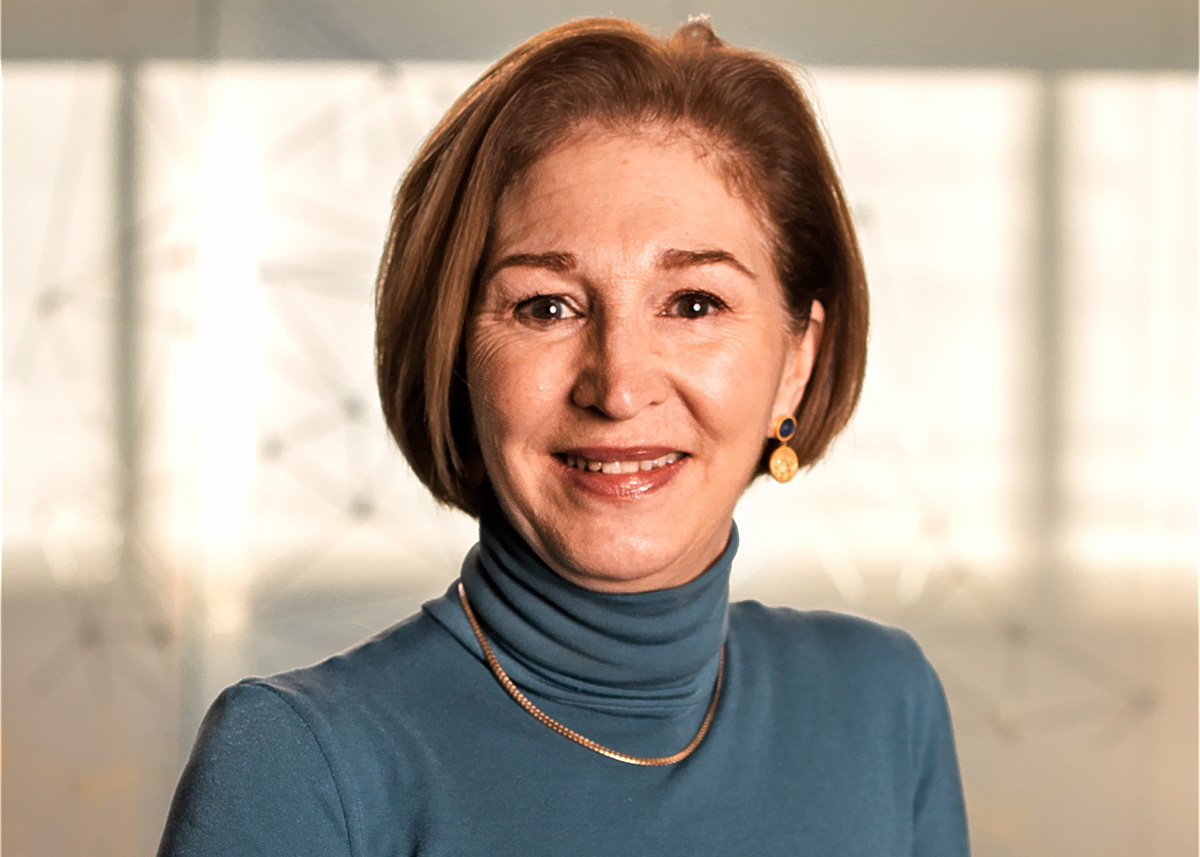 An individual with short brown hair looks toward the viewer.  They are wearing a blue turtleneck shirt and a gold necklace. 