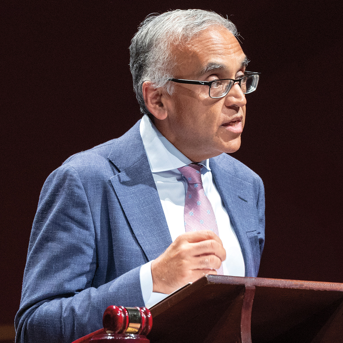 A photo of Ashish Jha, a person with light brown skin and gray hair. He is wearing glasses and a business suit and is speaking at a podium.