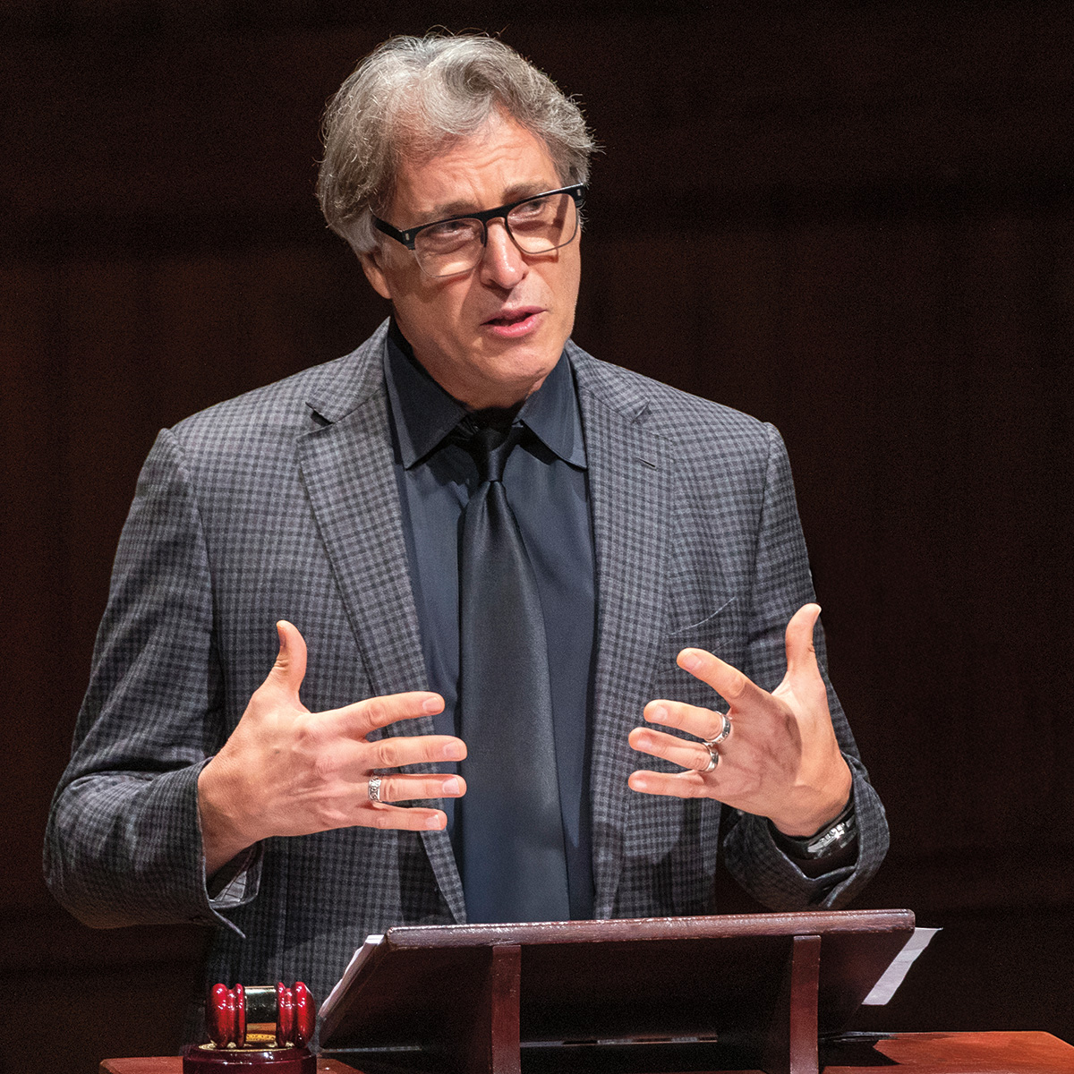 A photo of Brian Uzzi, a person with white skin and gray hair. He is wearing glasses and a business suit and is speaking at a podium.