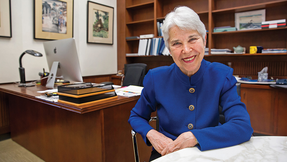 A photo of Carol T. Christ, a person with short straight white hair and pale skin. She sits at a desk in front of bookshelves, faces the camera, and smiles.