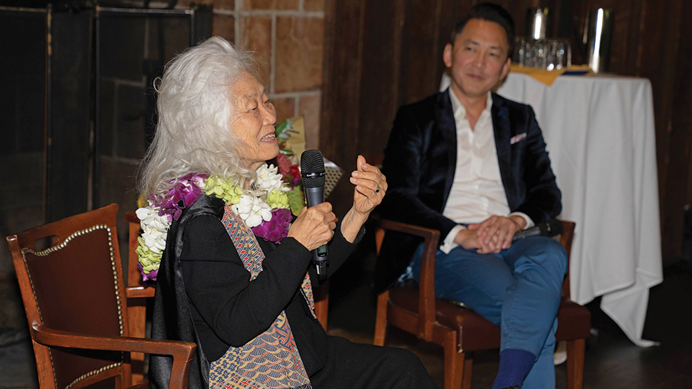 A photo of Maxine Hong Kingston, a person with brown skin and long wavy white hair. She wears a black dress under a multicolored scarf, and a necklace of white, green, and purple flowers. She sits and holds a microphone. Beside her is Viet Thanh Nguyen, a person with brown skin and short straight black hair. He wears a white collared shirt and a dark suit. He smiles at Kingston as she addresses the audience.