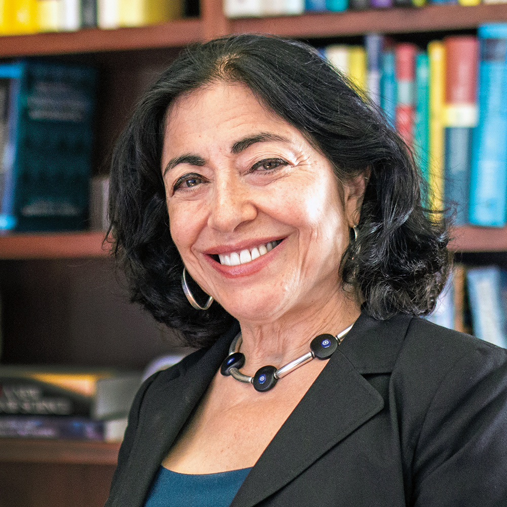 A woman is standing in a room with a bookshelf behind her. She is wearing a black suit and a blue shirt. She is smiling and appears to be posing for a picture.