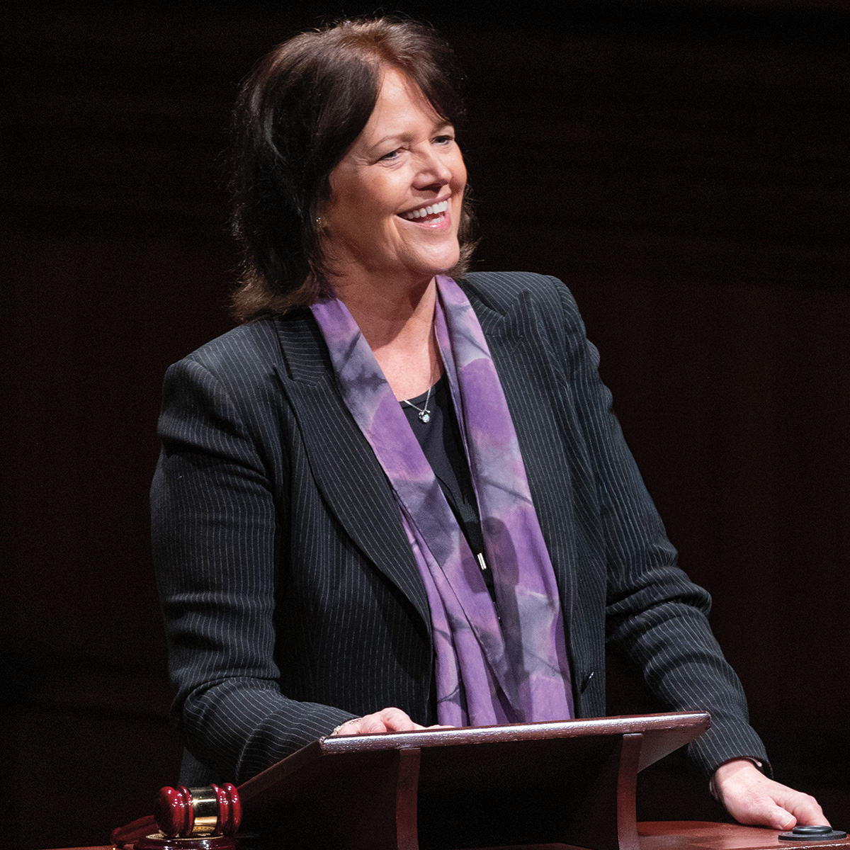 A photo of Christine Brennan, a person with light skin and medium-length brown hair. She is wearing a blazer and multicolored scarf and is speaking at a podium. 