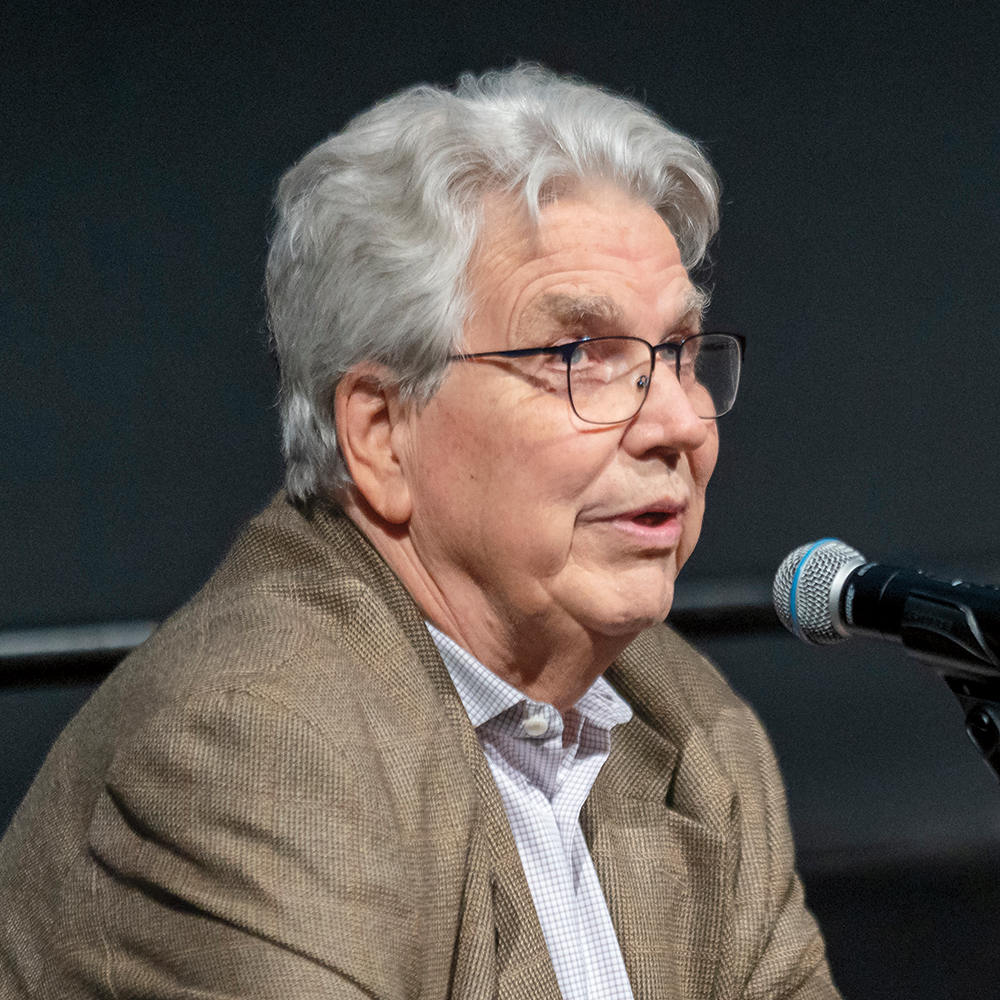 A man with gray hair and wearing wireframe glasses is sitting at a table and speaking into a microphone.