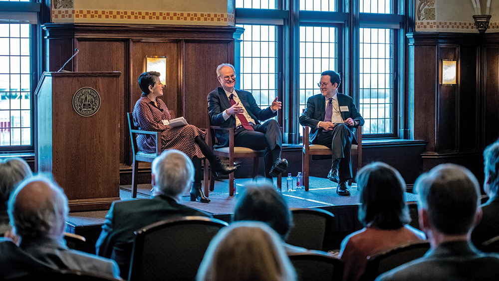 A photograph of three people seated on stage in front of an audience.