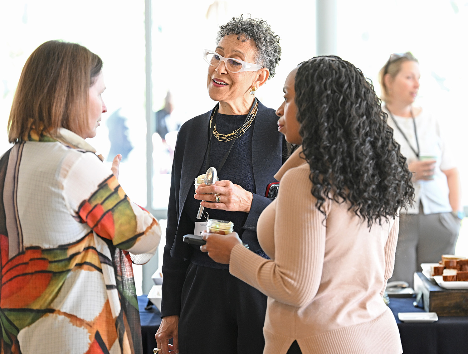 Three women are talking in a circle, holding snacks and beverages. 