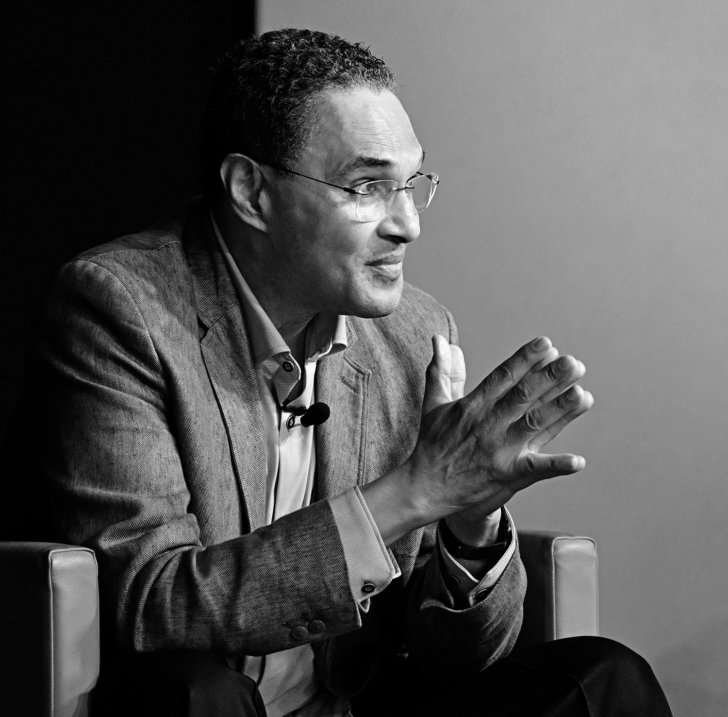 A man wearing glasses and a suit sits forward in his chair with his hands together. 