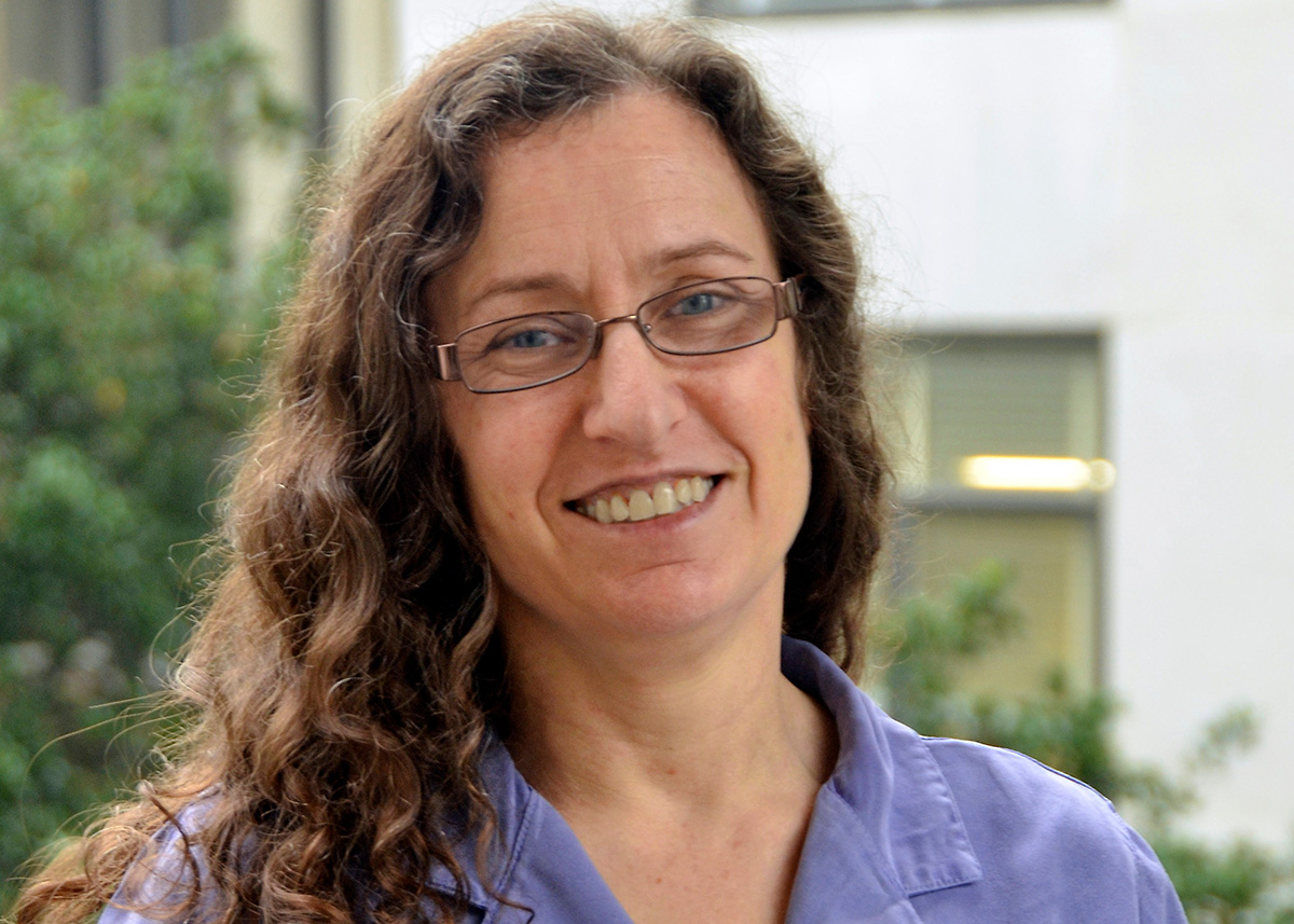 A photograph of a light-skinned individual with long curly brown hair. They are wearing glasses and smiling at the viewer. 