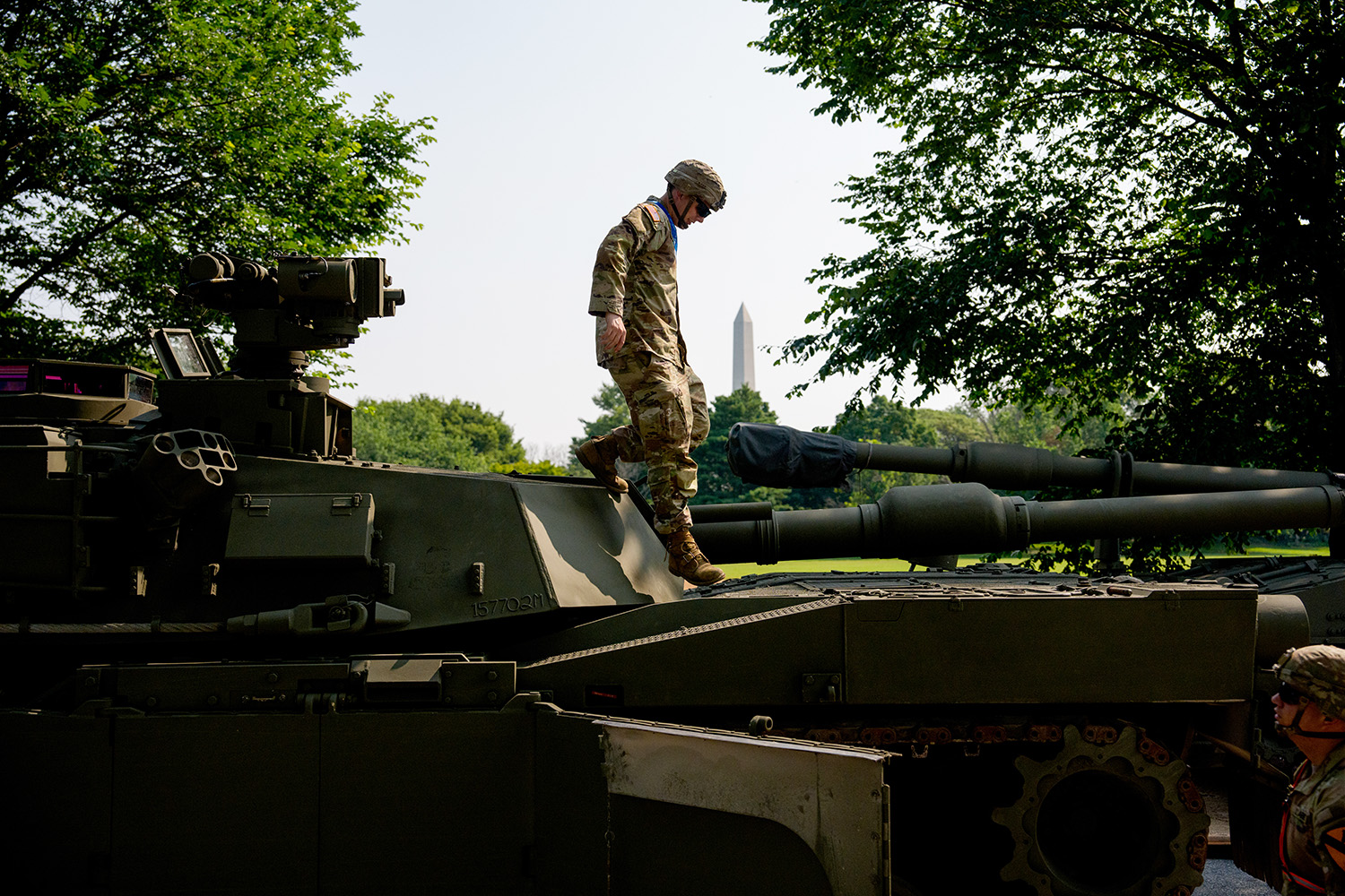 A U.S. Army soldier in uniform and headgear steps on top of an armored fighting vehicle. Around him are trees, a clear blue sky, and a view of the Washington Monument in Washington, D.C.