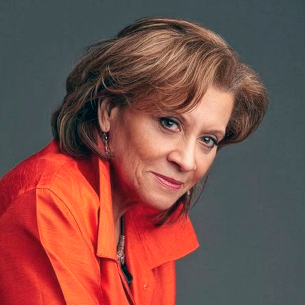 A person with short layered hair wearing an orange long-sleeve shirt is sitting with hands clasped on a gray background.