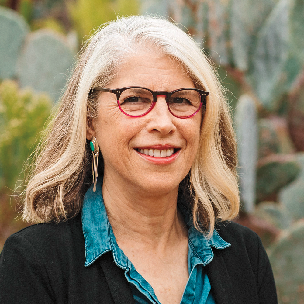A woman wearing glasses and a blue shirt is standing in front of a cactus plant.