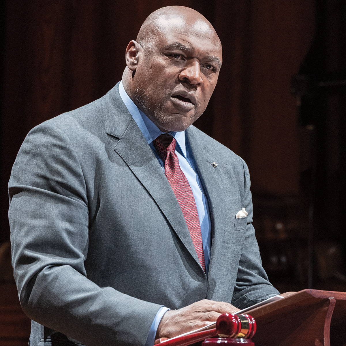 A photo of Gregory Robinson, a person with dark skin and a bald head. He is wearing a business suit and is speaking at a podium.