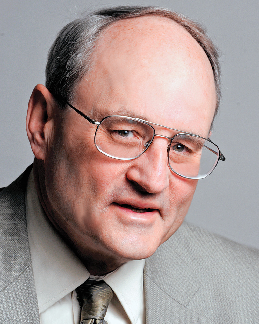 A headshot of Henry Brady wearing wire glasses and a gray suit. Brady has pale skin, blue eyes, and gray hair. He faces the viewer and smiles. 