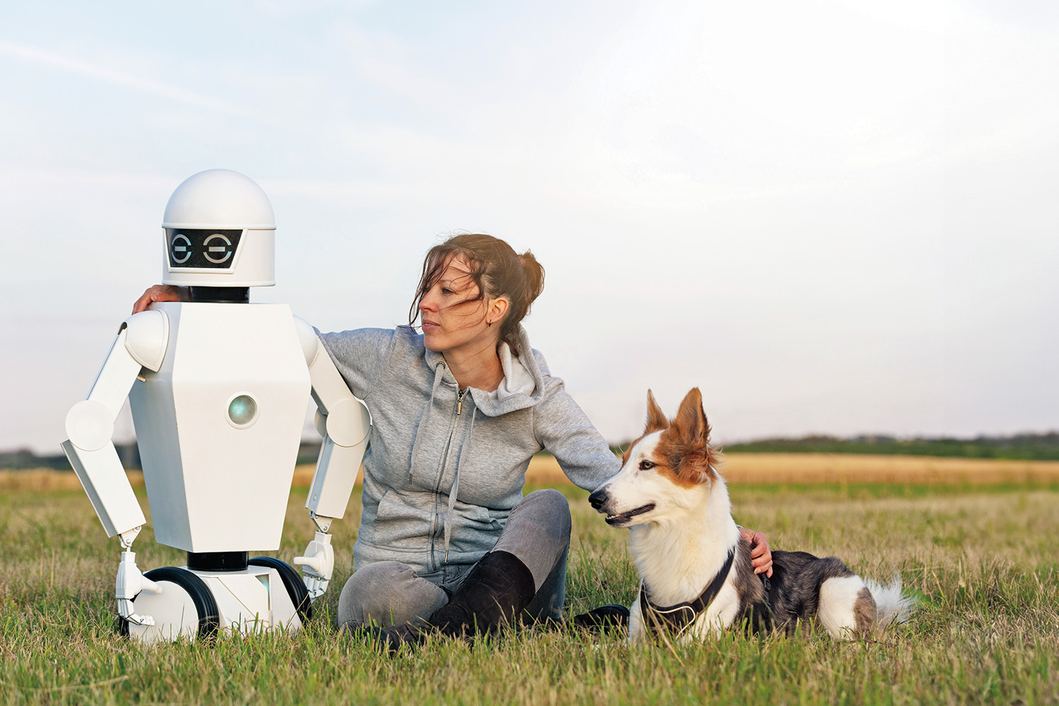 A person sitting in a field with their arms around a dog on their right and a robot on their left.