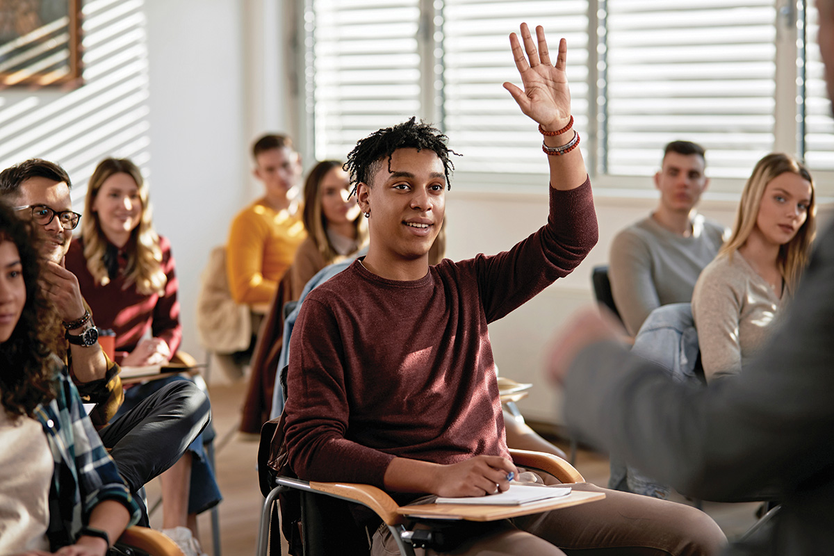 A person with short black curled locs and brown skin sits at a desk among a class of adult students. They raise their hand, smiling and face the instructor. 