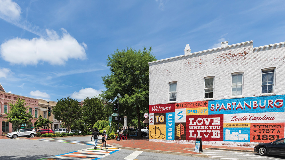 An intersection in a downtown area that has multiple building styles. A colorful mural can be seen in the foreground. Some of the mural reads “Spartanburg, South Carolina. Founded 1787, Inc. 1831.”