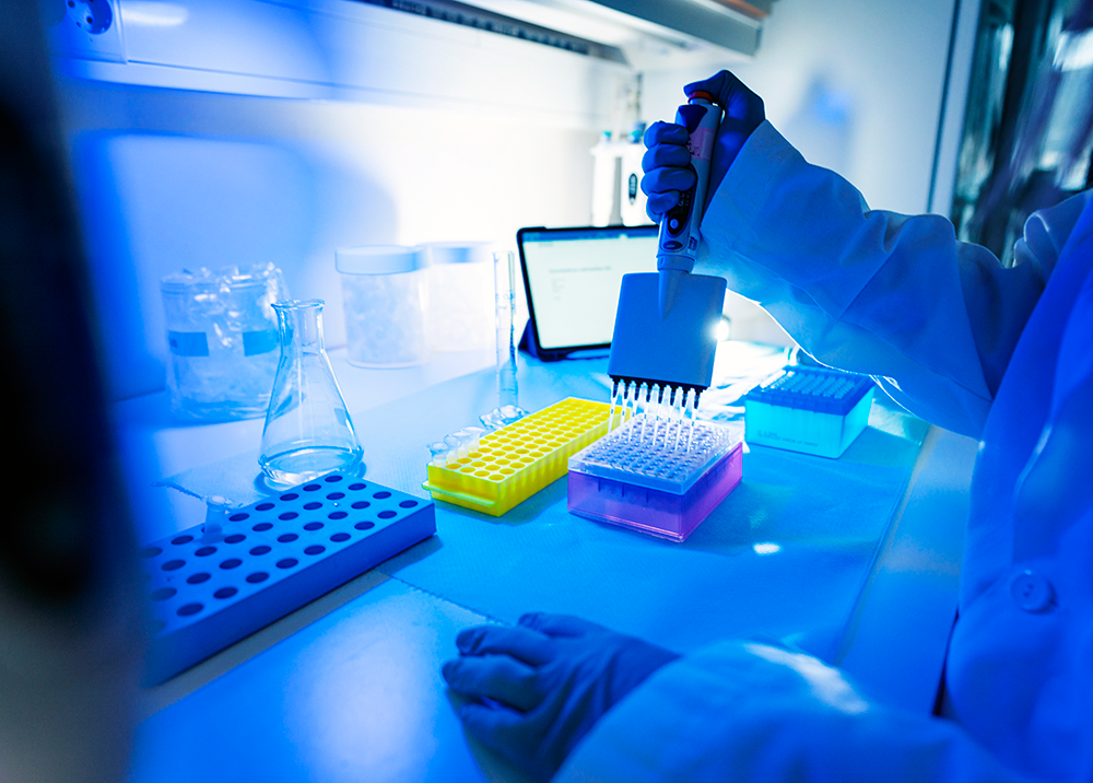 A scientist in a lab coat, wearing gloves, uses a multichannel pipette to transfer liquid into a well plate. Lab equipment and a computer tablet are on the workbench, and blue lighting illuminates the space.