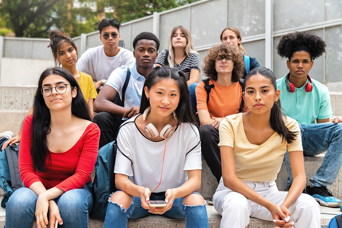 A group of young adults are sitting side by side on steps in an outdoor area. They are facing forward and looking at the camera.