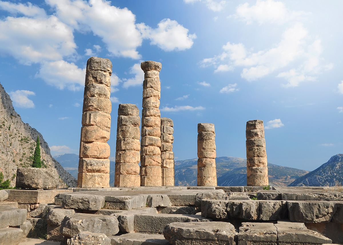 Ancient stone ruins with several standing columns of various heights overlook a mountainous landscape under a blue sky.