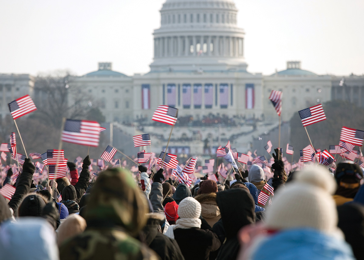 A view facing the back of crowd of people near the Capitol Building in Washington, D.C. Many people in the crowd wave small American flags. 