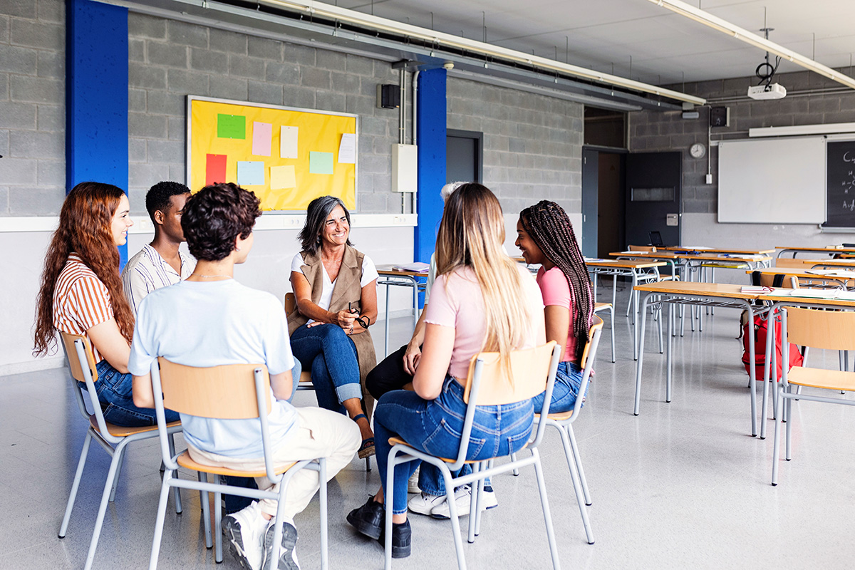 Seven people sitting in chairs in a circle in a classroom setting. Their attention is focused on one member of the group, who is speaking. 