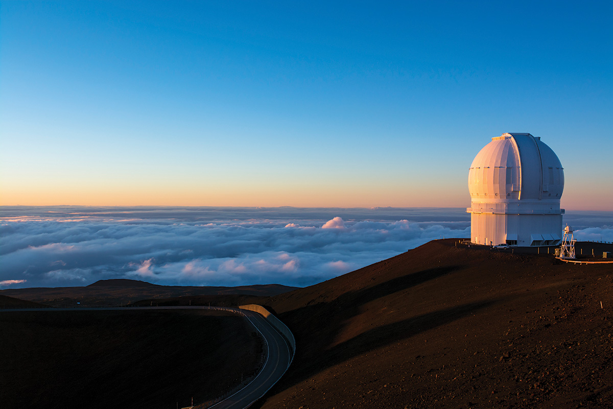 An aerial view of a road beside a planetarium on a mountainside. Clouds hover in the background, obscuring the view of the ground below.