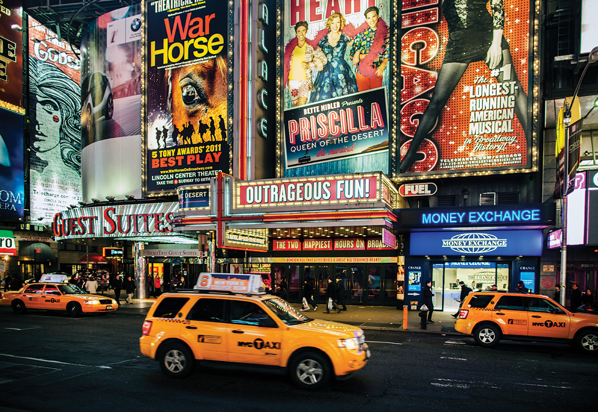Ads for Broadway shows light up above taxis in Times Square at night.  