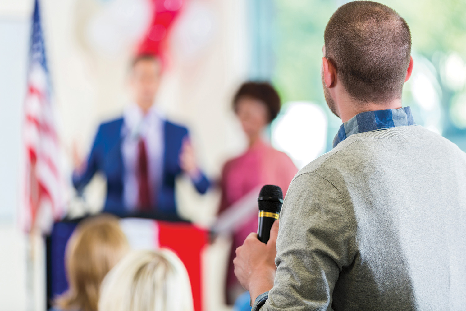 A man standing in the audience with a microphone in his hand asks a question of a man and woman standing at a podium in front of him. 