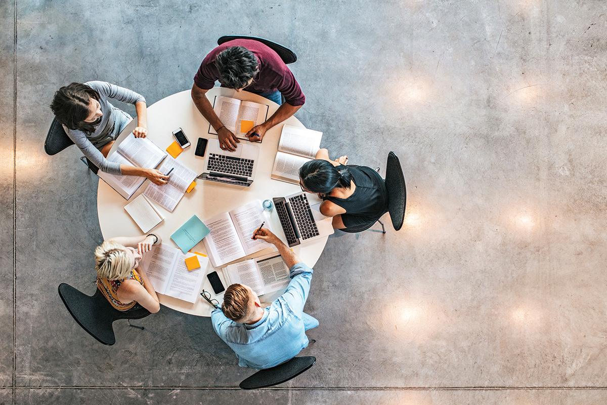 An aerial view of five people sitting at a round table with open books and laptops between them. They take notes and lean over their materials to engage with each other.