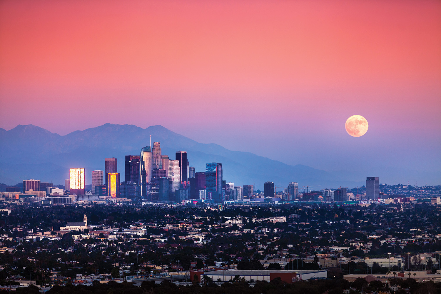 A sunset in hues of pink above the city skyline of Los Angeles, with skyscrapers in the background and residential areas and trees in the foreground. A full moon hangs low in the sky to the right.