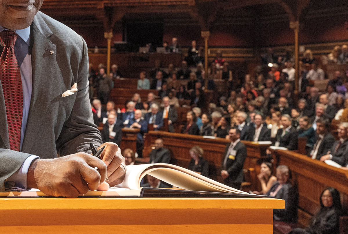 A person, wearing a business suit and tie, is writing in a book with white pages. He is holding a pen in each hand. We see part of his face, from his lips down. In the background is an audience seated in a large auditorium.