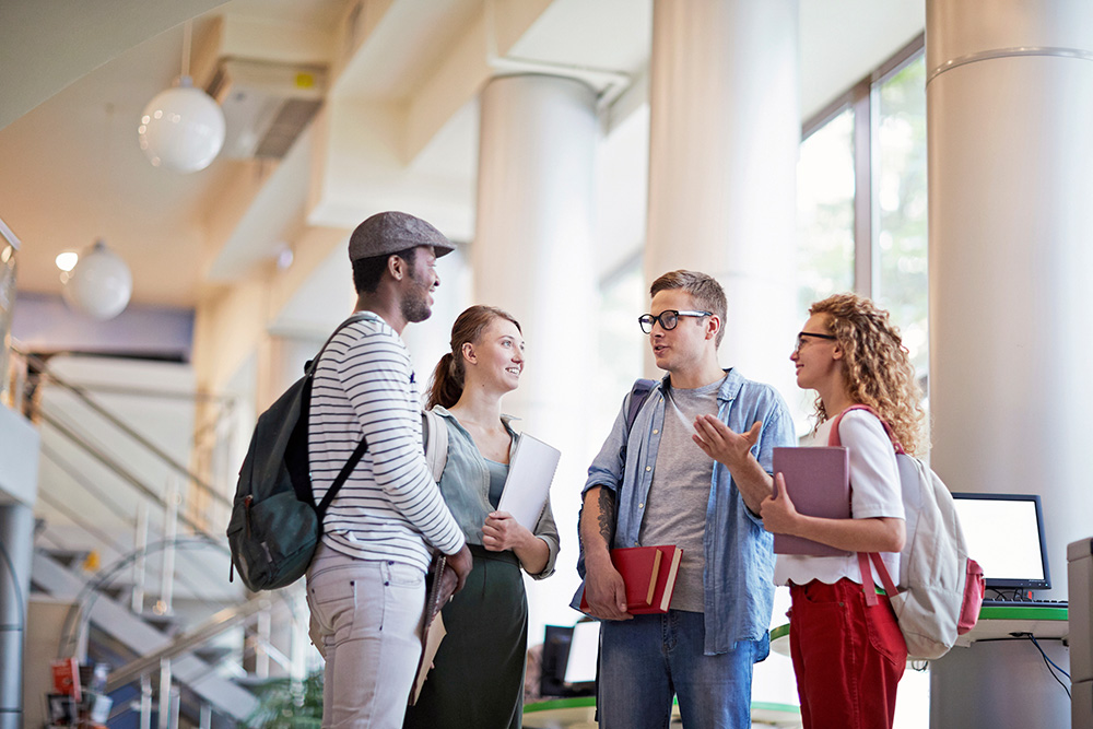 Students are seen standing in a university building engaged in casual conversation while holding their schoolbooks and belongings.
