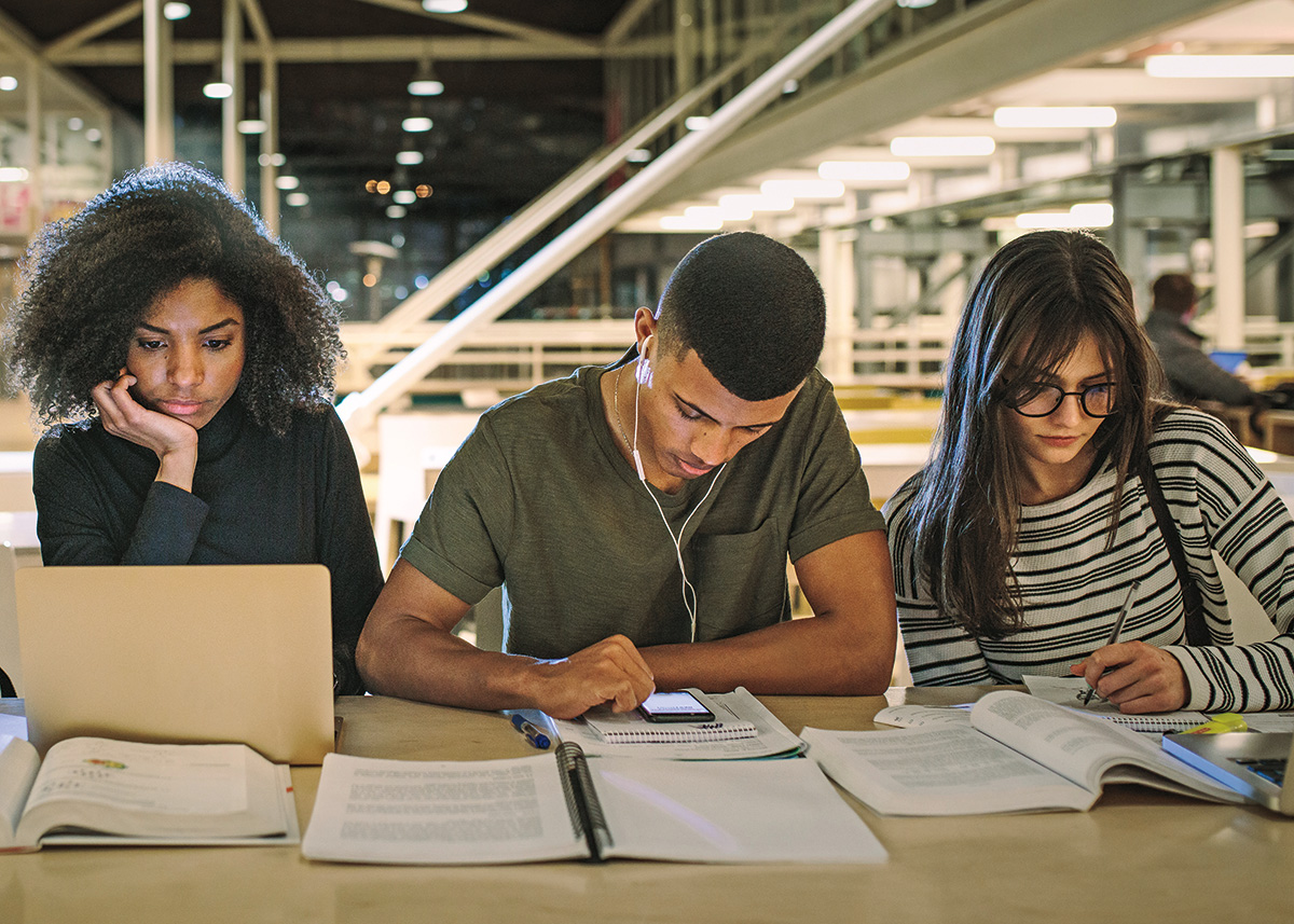 Four college-age students study together at a table covered by their laptops, phones, and books. Two students are engaged on their phones, another student looks at their laptop, and the last student is writing in a notebook.