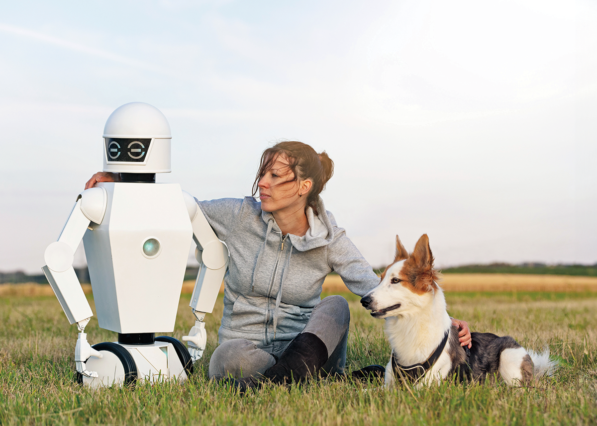 A person sitting in a field with their arms around a dog on their right and a robot on their left.