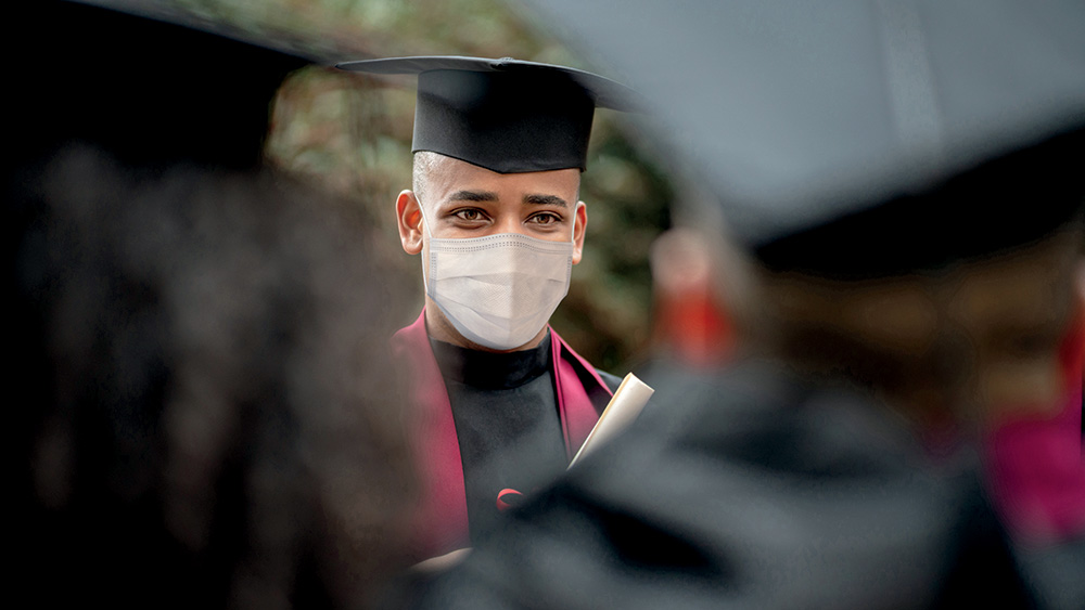 A person with light brown skin and a shaved head wears a graduation cap and gown, as well as a surgical mask. They face their fellow graduates. 