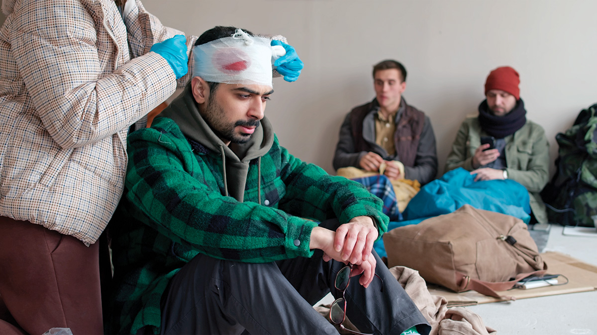 An adult sits on the floor beside a backpack. They have pale skin, a thick dark mustache, and short black hair. A person stands behind them and bandages the top of their head. Red can be seen through the bandages. Two other people wait in the background.