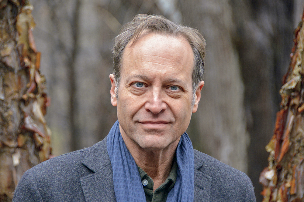A headshot of Jeffrey Brown. Brown has pale skin, blue eyes, and graying dark hair. He wears a blue scarf, a gray coat, and smiles at the viewer. 