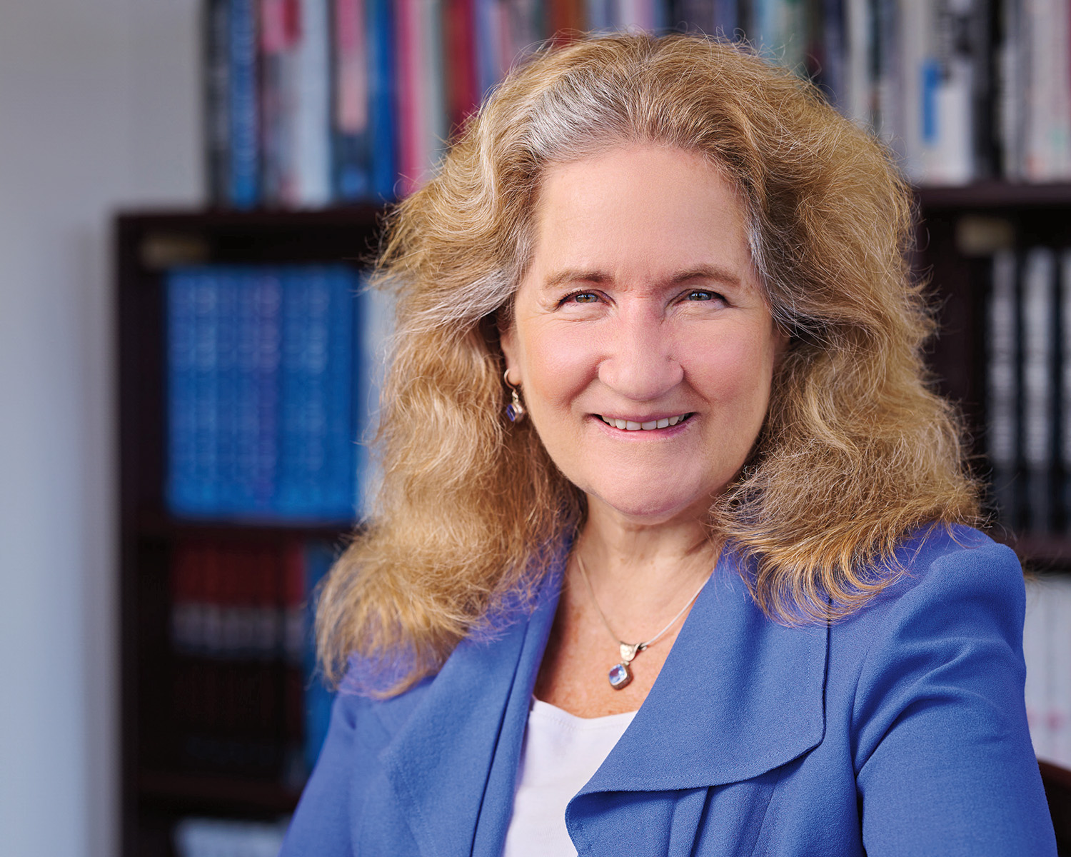 A headshot of Katherine S. Newman, a person with long gray and brown hair and pale skin. Newman is wearing a bright blue blazer and white blouse and smiling at the viewer.