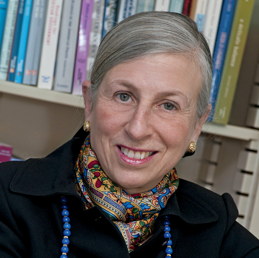 A headshot of Kay Lehman Schlozman wearing a multicolored paisley scarf, a blue necklace, and a black suit. Schlozman has pale skin, blue eyes, and long gray hair. She faces the viewer and smiles. 