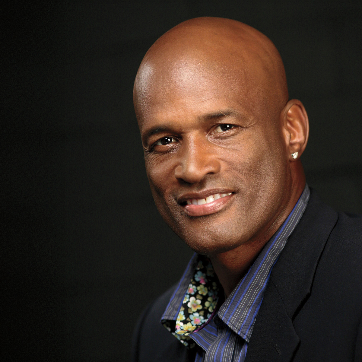 A photo of Kenny Leon, a person with dark skin and a bald head, who is wearing a dark suit jacket, a blue striped shirt with a flower pattern on the inside collar, and an earring. He is in front of a dark background and is smiling at the viewer.