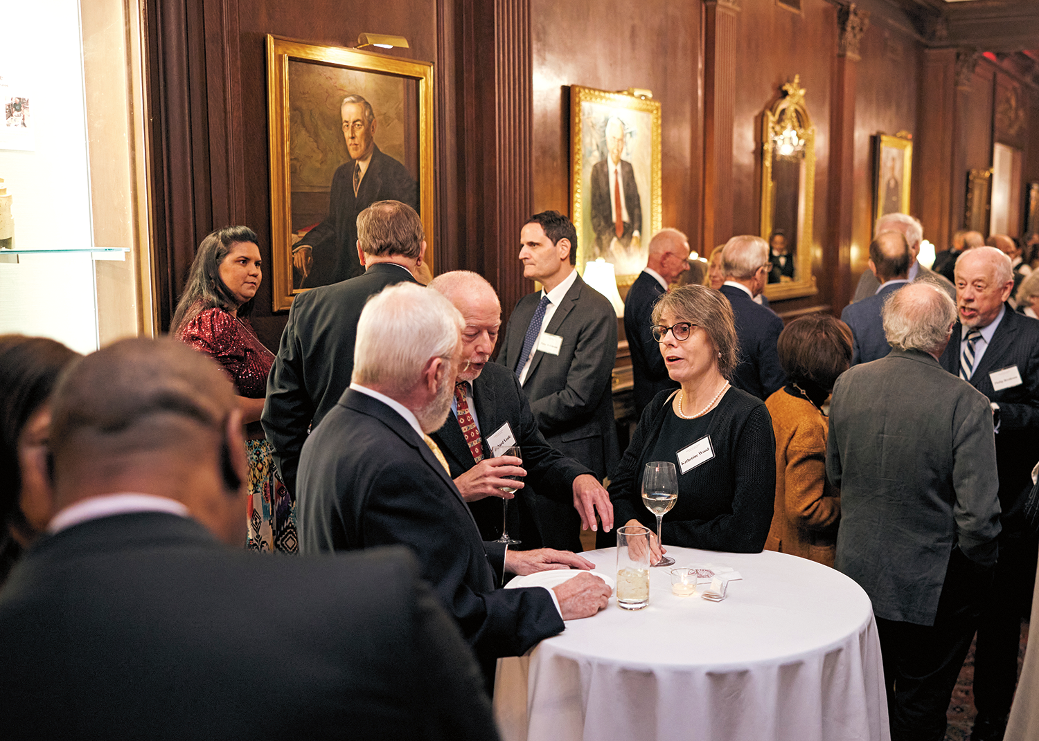 A candid photo of formally dressed adults at a reception in a room with wood paneling and gold framed portraits.