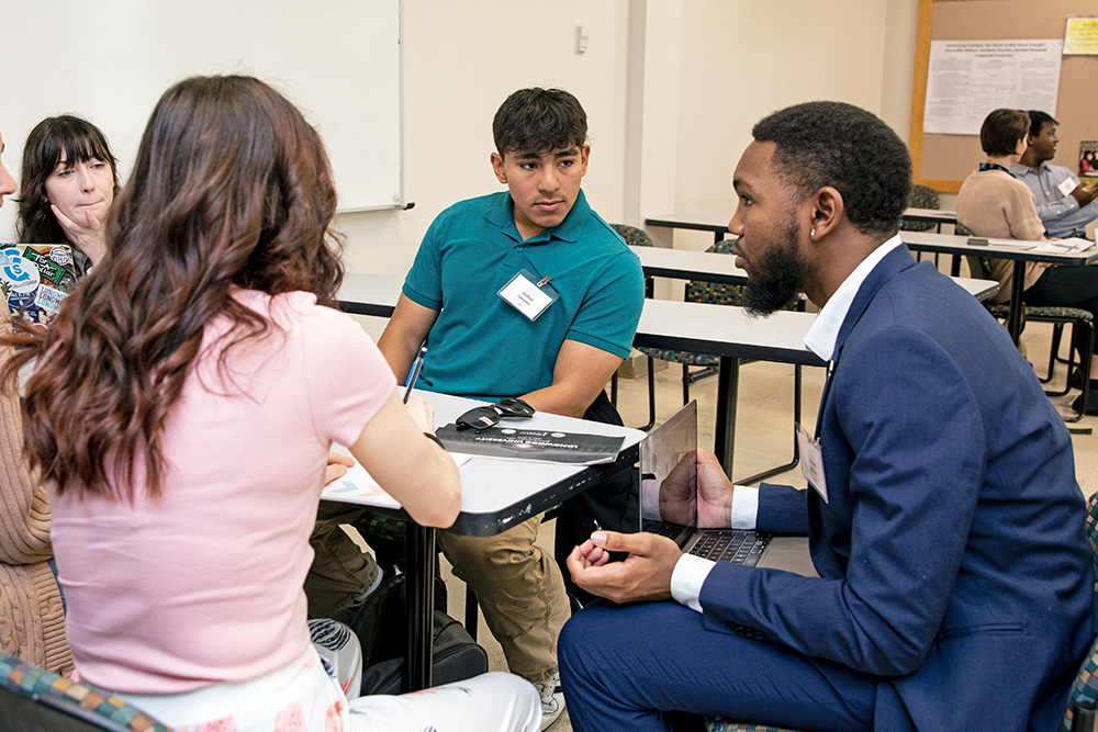 A faculty member in a suit addresses a few students sitting around a classroom table.