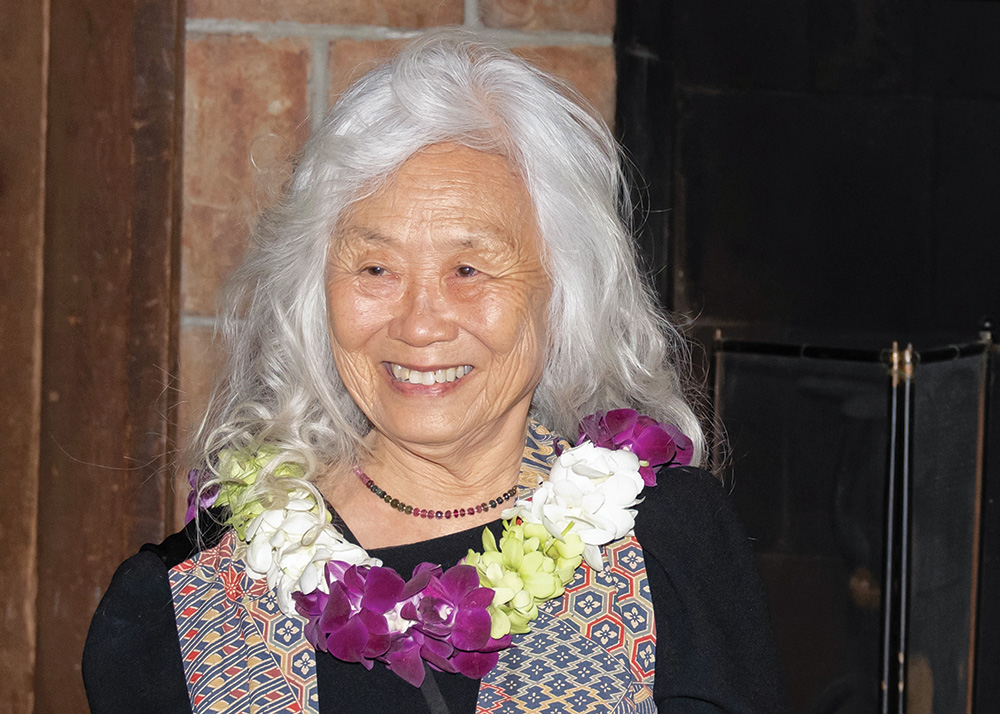 A photo of Maxine Hong Kingston, a person with brown skin and long wavy white hair. She wears a black dress under a multicolored scarf, and a necklace of white, green, and purple flowers. She looks to her right and smiles.