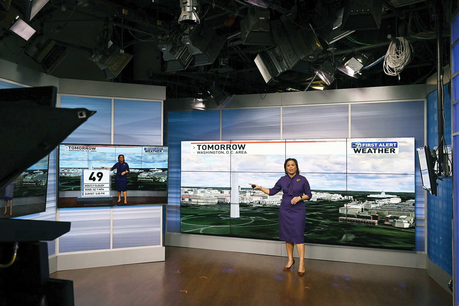 A photo of a television studio shows a camera in the foreground and a meteorologist in the background gesturing toward a monitor where a landscape view of Washington D.C. is displayed. The meteorologist has straight brown hair and brown skin, and wears a purple dress and brown heels. A smaller monitor is a few feet away, which shows a weather forecast along the bottom of the screen. The rest of the screen mirrors the image of the meteorologist.
