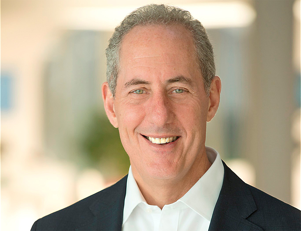 A light-skinned individual with short gray hair smiles and looks toward the viewer.  They are wearing a white business shirt and dark blazer. 