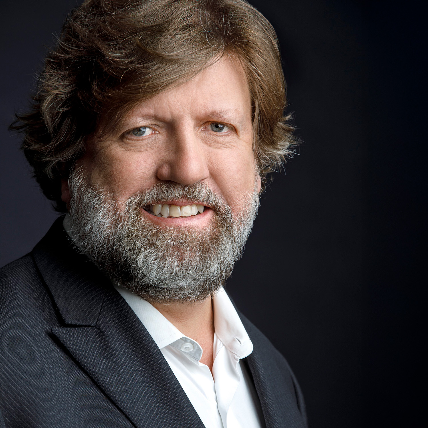 Oskar Eustis is smiling at the viewer. He has medium-length brown hair and a gray and brown beard and mustache. He is wearing a dark business jacket and white shirt.